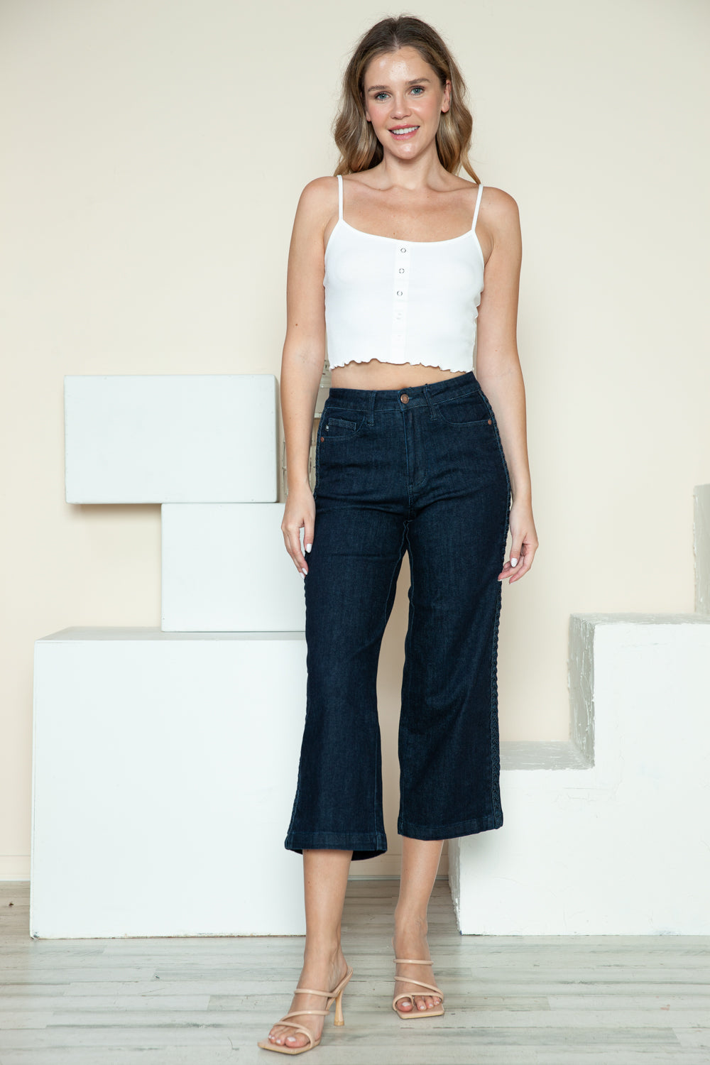 Person wearing Judy Blue Full Size Side Seam Braid Detail Crop Wide Leg Jeans and beige strappy heels, standing on a wooden floor with a white background.