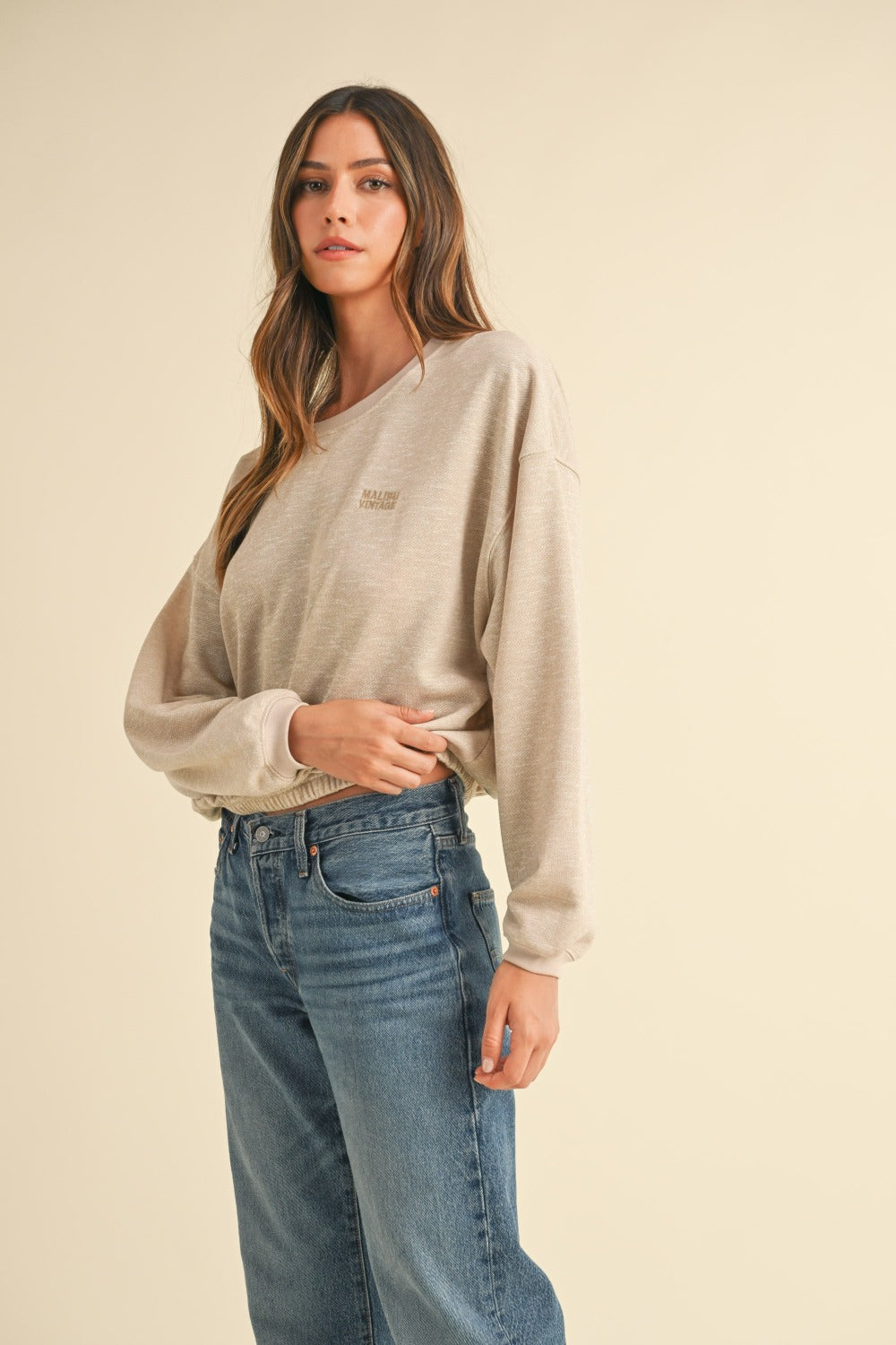 A woman with long brown hair wears the Mable Round Neck Letter Embroidery Crop Sweatshirt in beige, paired with blue jeans, standing confidently with one hand in her pocket against a plain background, perfectly capturing effortless street style.