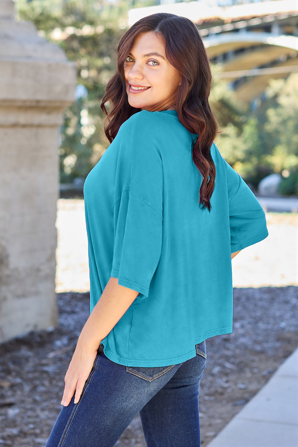 A person with long dark hair and a bright smile stands outdoors, showcasing their basic style in a loose, slightly stretchy *Basic Bae Full Size Round Neck Drop Shoulder T-Shirt* in pink and blue jeans, with one hand on their hip.