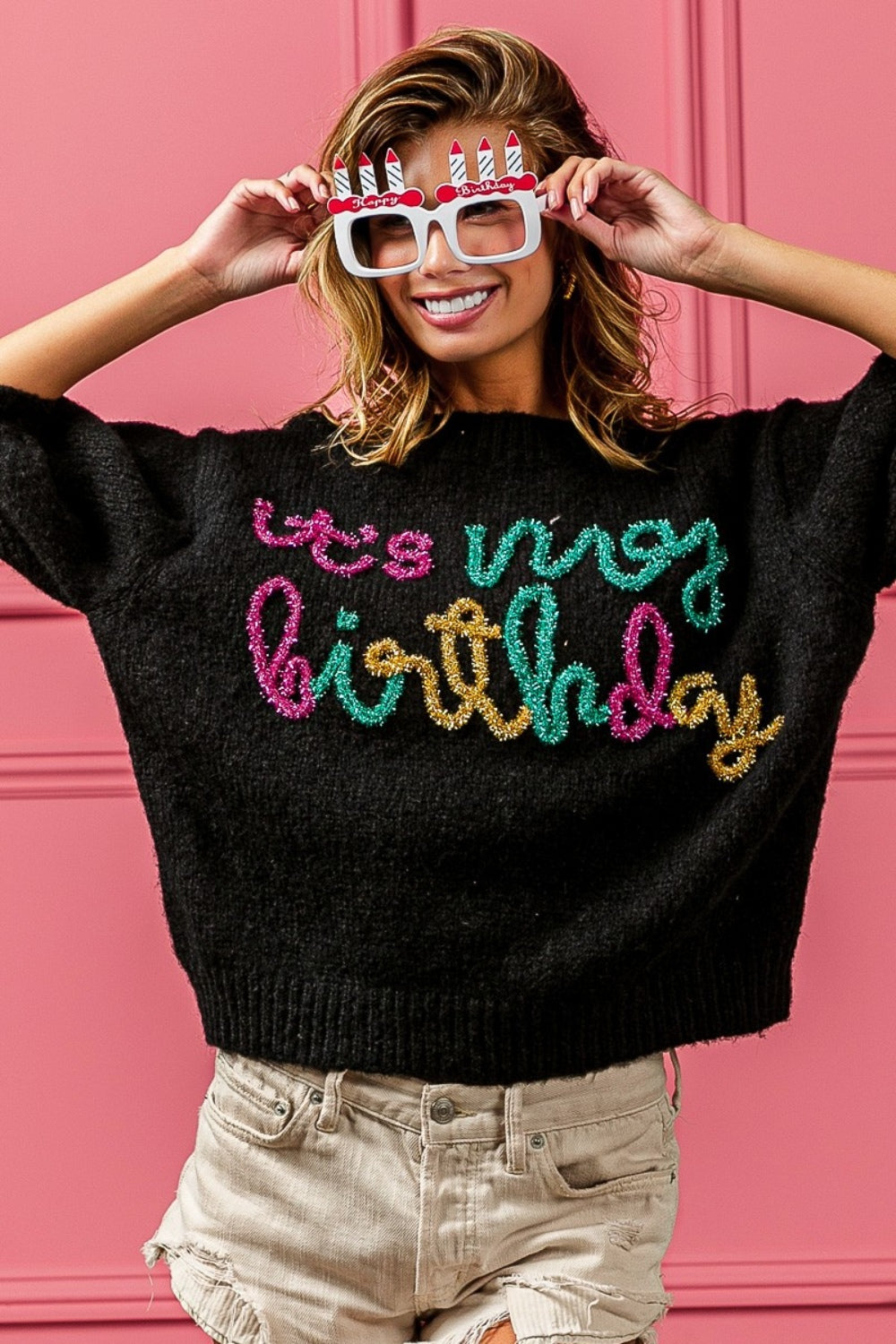 A woman poses against a pink wall, wearing a BiBi Metallic Letter Puff Sleeve Hairy Sweater that reads "It's My Birthday," beige shorts, and novelty glasses with birthday candles.