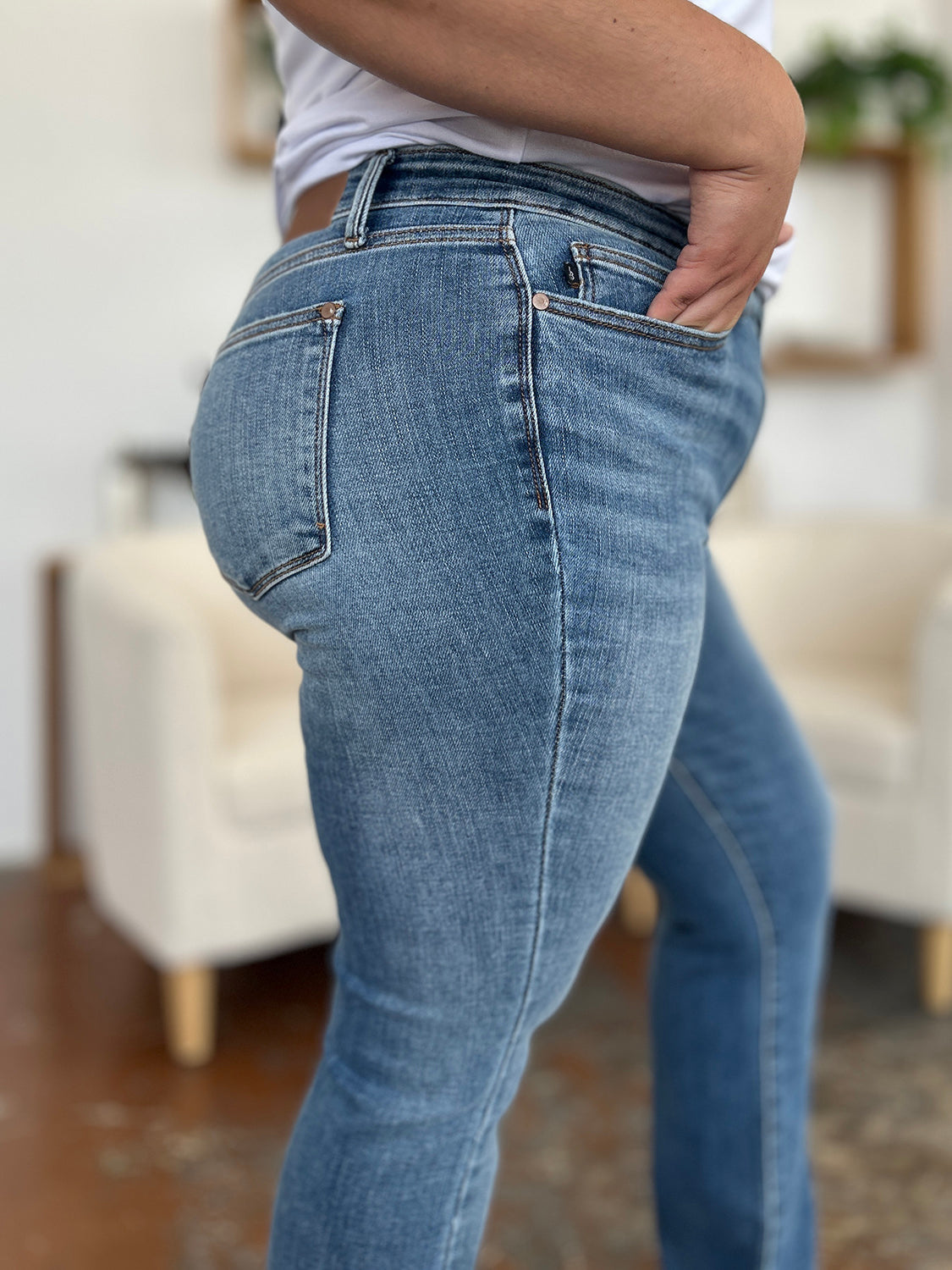 Person wearing Judy Blue Full Size Mid-Rise Waist Straight Jeans and a black top, standing in a room with white chairs, potted plants, and wooden shelves. Only the lower half of the body and part of the arms are visible.