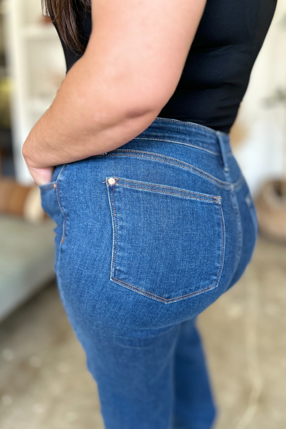 A person wearing Judy Blue Full Size High Rise Straight Jeans, a black top, and white shoes stands gracefully on a polished concrete floor.