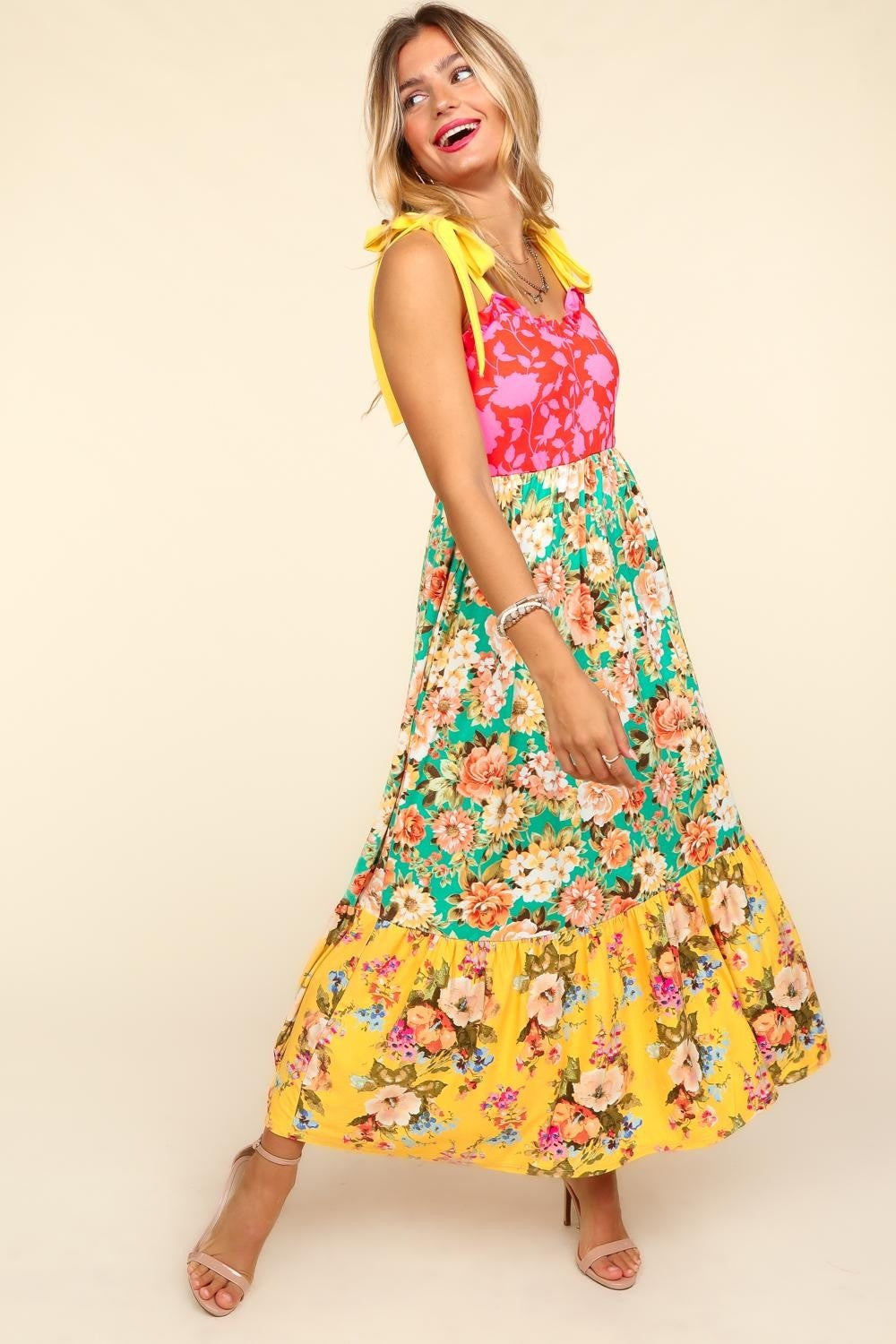 A person wearing the Haptics Floral Color Block Maxi Dress with Pockets, featuring a vibrant floral print and a color block maxi skirt in pink and yellow hues, stands smiling against a beige background.