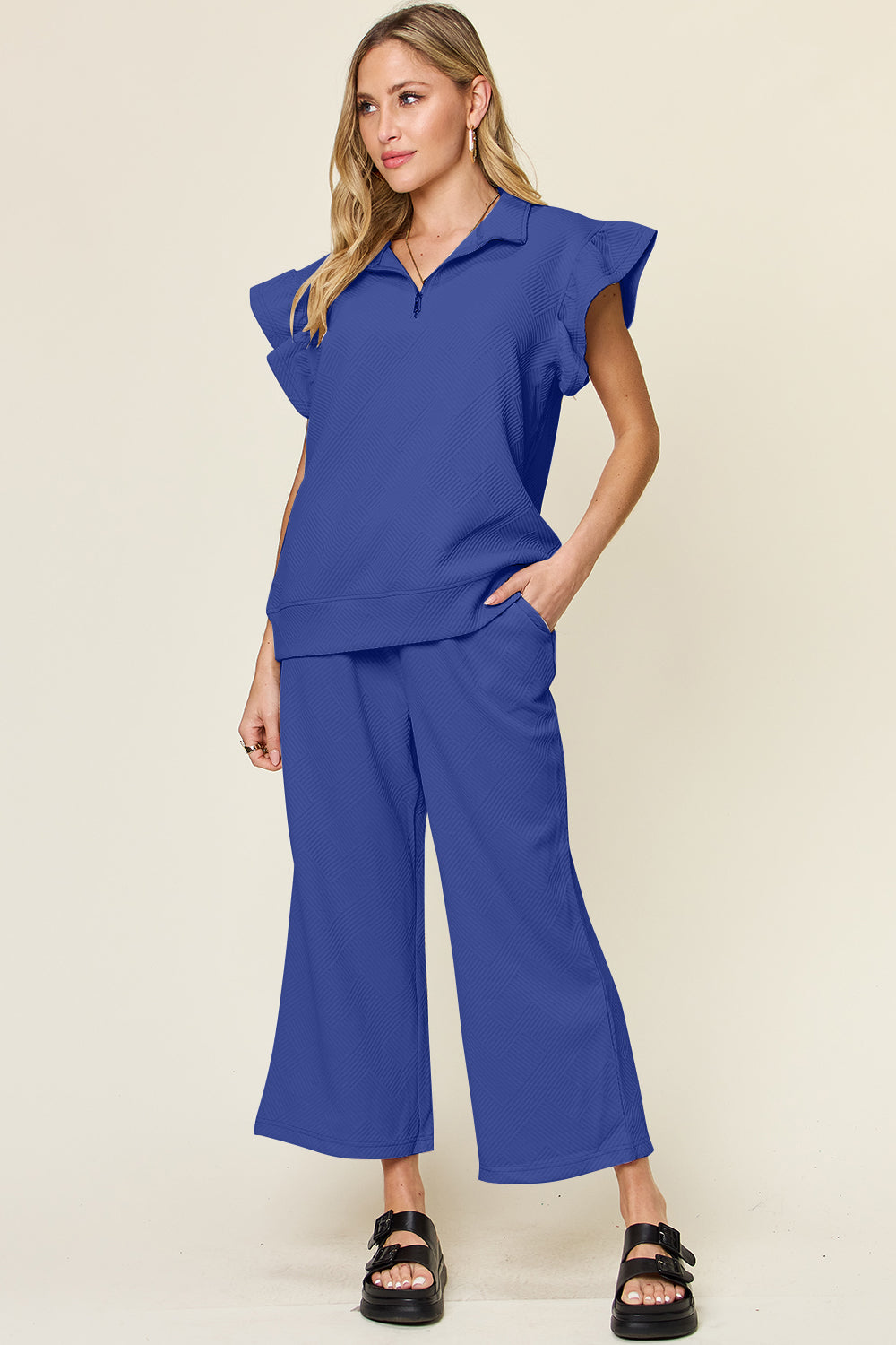 A woman stands wearing the Double Take Texture Ruffle Short Sleeve Top and Drawstring Wide Leg Pants Set in a slightly stretchy, light gray fabric with zipper detail. The two-piece outfit is complemented by black sandals, and she has one hand in her pocket. Machine wash cold for easy care.