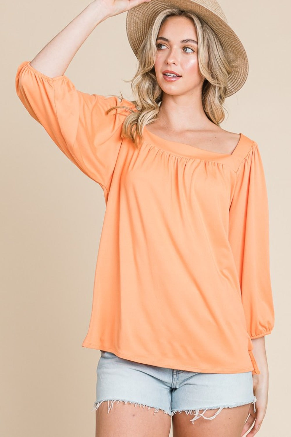 A person with long blonde hair is smiling and wearing the trendy Culture Code Square Neck Puff Sleeve Top in a light orange hue, paired with light denim shorts, showcasing a versatile fashion piece against a beige background.