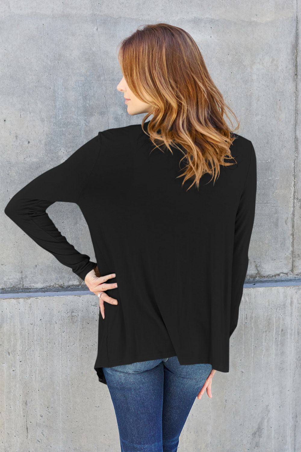 A woman wearing the Basic Bae Full Size Open Front Long Sleeve Cardigan in navy blue, paired with a white top and blue jeans, stands against a concrete wall. She touches her hair and gazes to the side, embodying a chic yet simple style. The slightly stretchy materials of the cardigan ensure comfort while maintaining its stylish simplicity. For easy care, machine wash it cold.