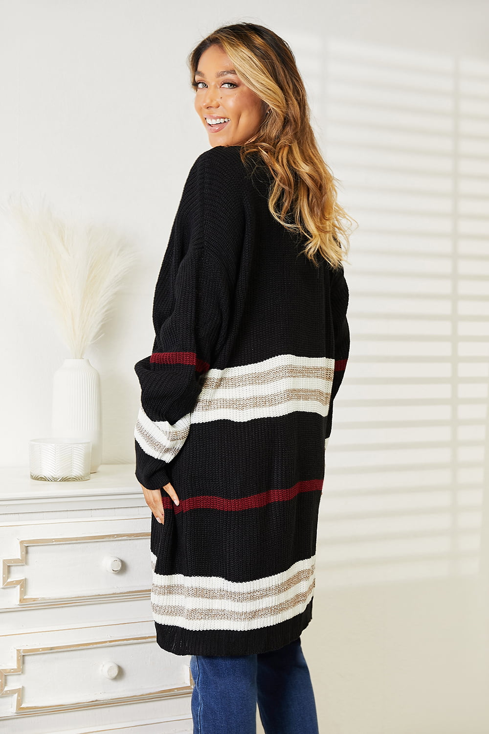 A person wearing a Perfee Striped Rib-Knit Drop Shoulder Open Front Cardigan with white and red accents, a white top, blue jeans, and layered necklaces stands next to a decorative table.