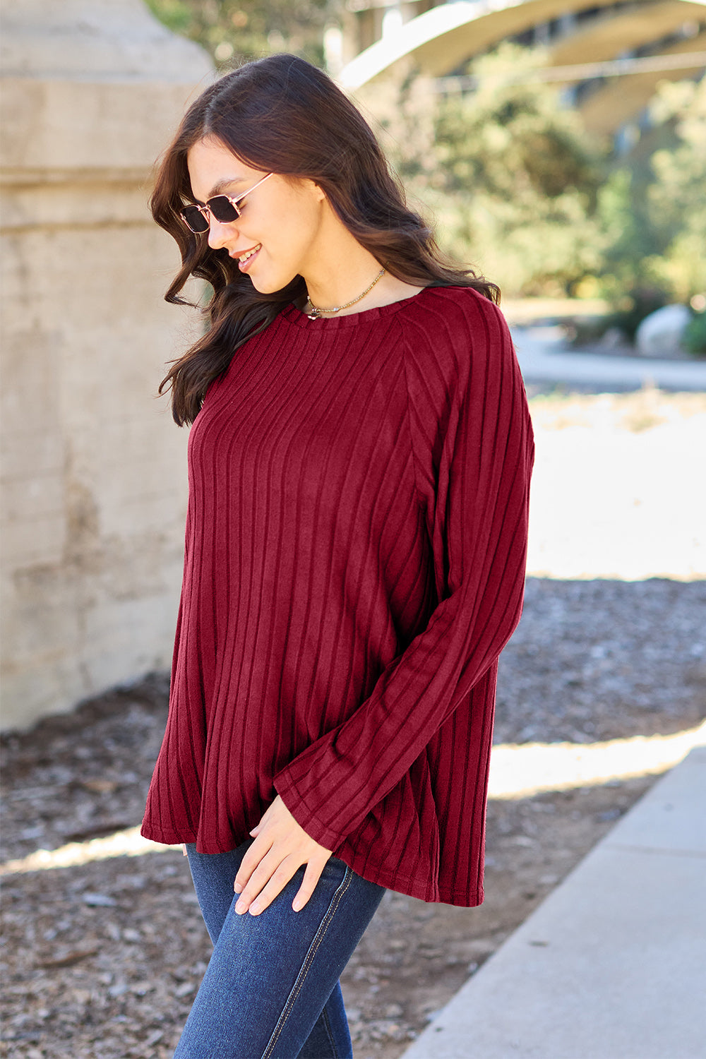 A woman with long brown hair wearing sunglasses and a Basic Bae full-size, slightly stretchy ribbed round neck long sleeve knit top in red, paired with blue jeans, is standing outdoors.