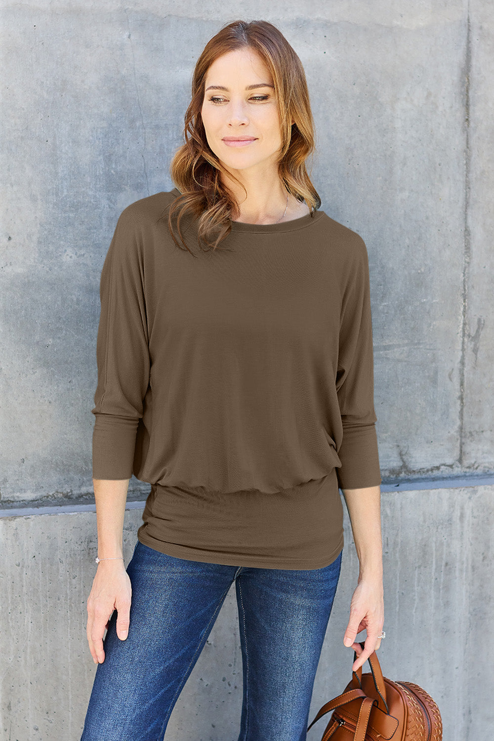 A woman in a slightly stretchy, brown Basic Bae Full Size Round Neck Batwing Sleeve top and blue jeans stands against a concrete wall, holding a brown handbag.