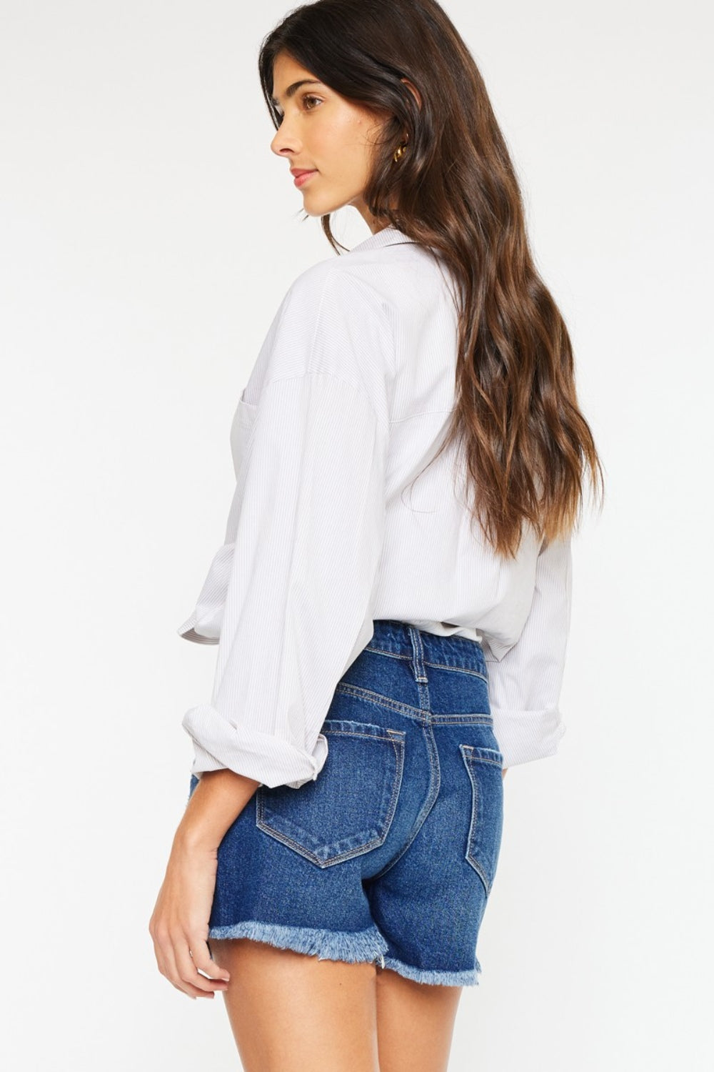 A woman with long dark hair wears a white blouse tied at the waist over a white top and Kancan High Rise Frayed Hem Denim Shorts, posing with one hand in her pocket against a plain background, embodying the perfect summer wardrobe.