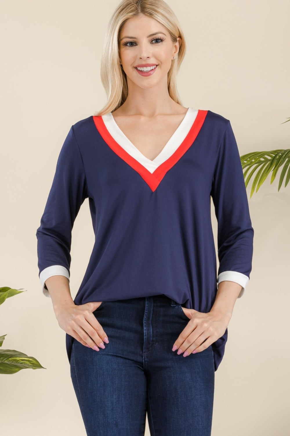 A woman with long blonde hair wearing the Celeste Full Size V-Neck Contrast T-Shirt crafted from lightweight material and dark blue jeans stands smiling.