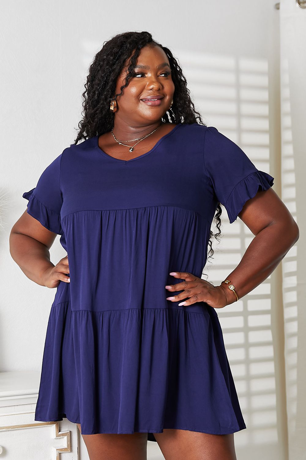 A woman stands indoors wearing the Mandy V-Neck Flounce Sleeve Tiered Dress in navy blue. She has one hand on her hip and is smiling.