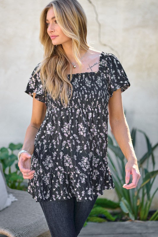 A woman stands outdoors donning a Floral Printed V-Neck Ruffle Top paired with jeans, set against a neutral background, with plants in the foreground, highlighting her fashion-forward wardrobe.