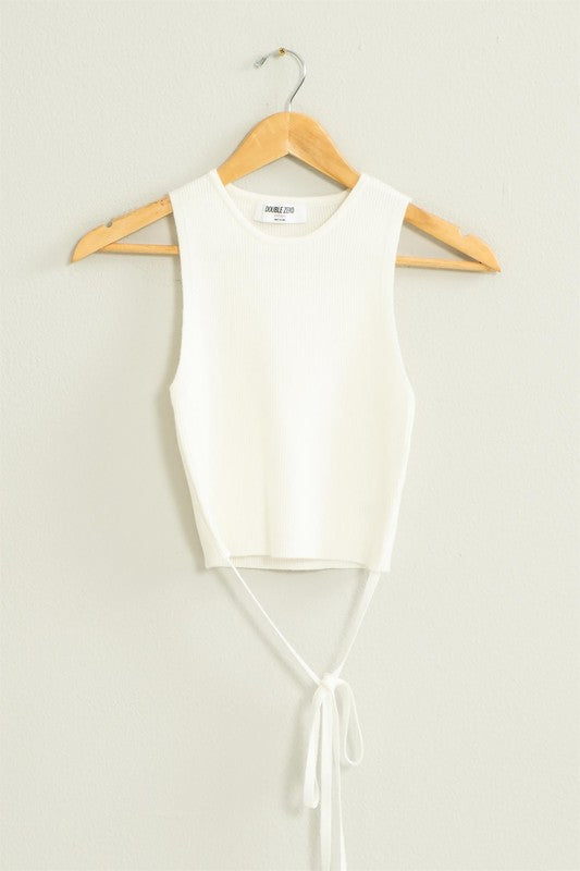 The Good Times Ribbed Sweater Tank Top, featuring a light green color, cropped hem, and back tie, hangs on a wooden hanger against a beige wall.