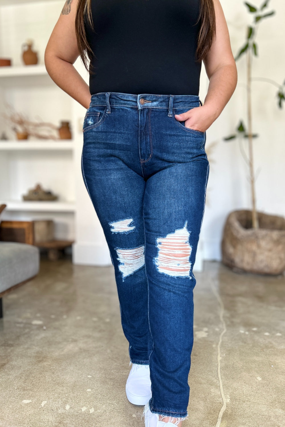 A woman in a black sleeveless top and Judy Blue Full Size High Waist Rigid Magic Heavy Destroy Straight Jeans stands indoors with her hands in her pockets. The background features a plant and shelves adorned with decor items.