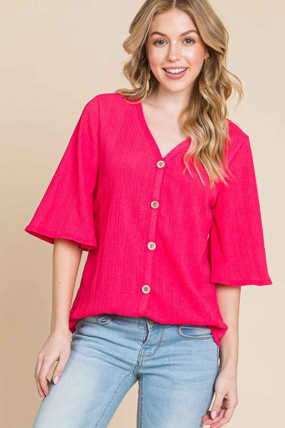 A person is wearing the BOMBOM Texture Decorative Button V-Neck Top, a chic design in bright pink featuring elegant details like short bell sleeves, and pairing it with blue jeans, stands against a beige background.