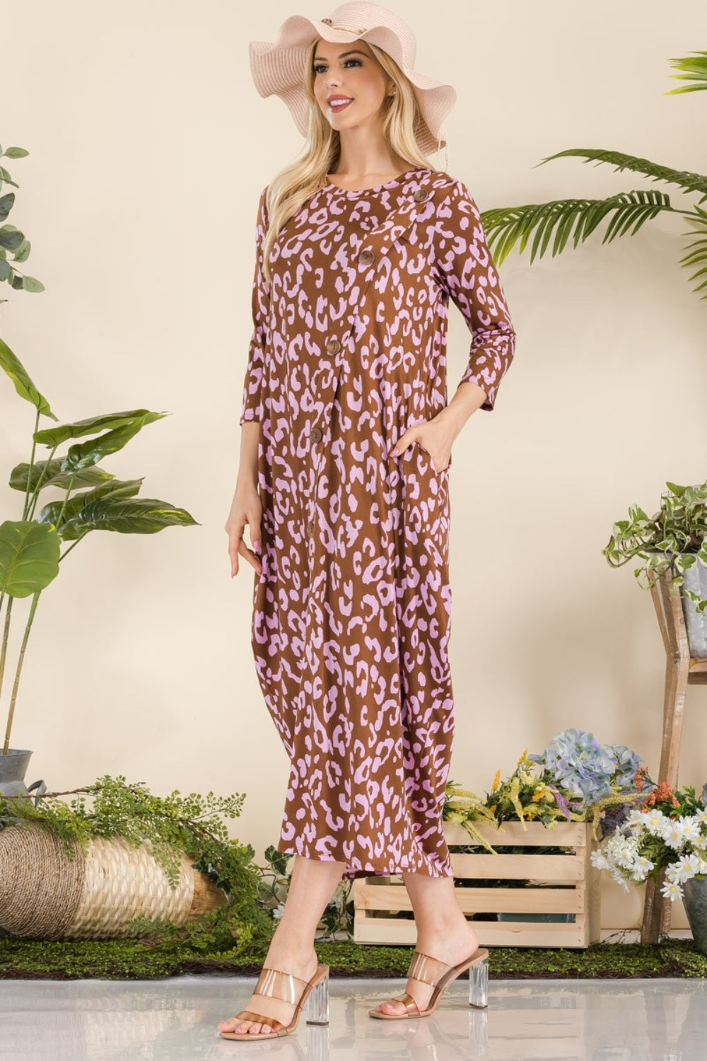 A woman in the Celeste Full Size Leopard Contrast Dress with pockets, featuring a brown and pink leopard print design and 3/4 sleeves, stands smiling among potted plants and wooden crates while wearing a wide-brim hat and sandals.