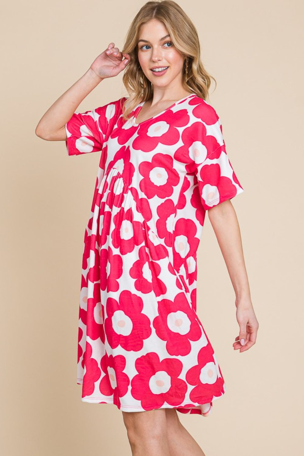 A woman gracefully stands against a beige background, wearing the BOMBOM Flower Print Ruched Dress adorned with large pink blooms. The stretchy fabric of the dress complements her form, effortlessly enhancing its elegant silhouette.