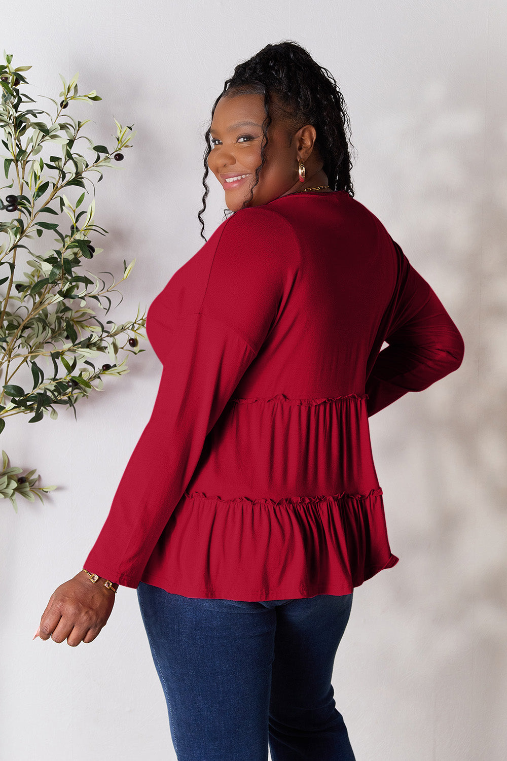 Person stands indoors near a plant, wearing the Double Take Half Button Long Sleeve Ruffle Hem Blouse in gray and blue jeans, smiling while looking to the side. The slightly stretchy fabric of the blouse adds comfort while its buttoned design offers a touch of elegance.