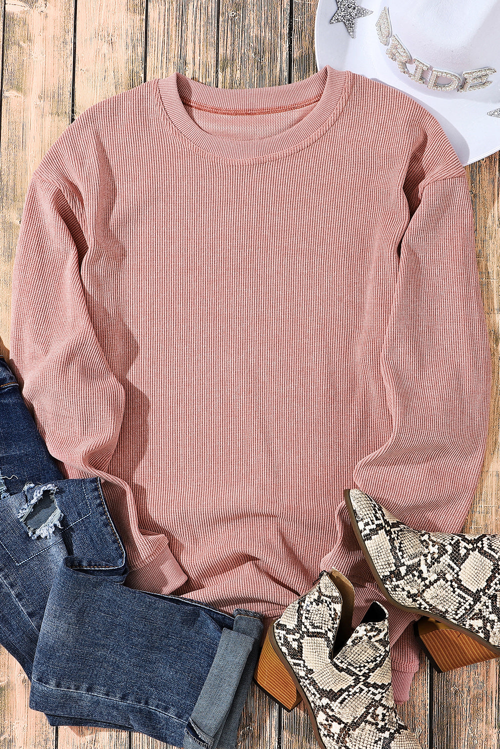 A woman with long, wavy hair is sitting on a high stool, smiling and holding a tumbler. She is wearing a relaxed fit Pink Solid Ribbed Knit Round Neck Pullover Sweatshirt, white shorts, a chain necklace, and knee-high white boots.