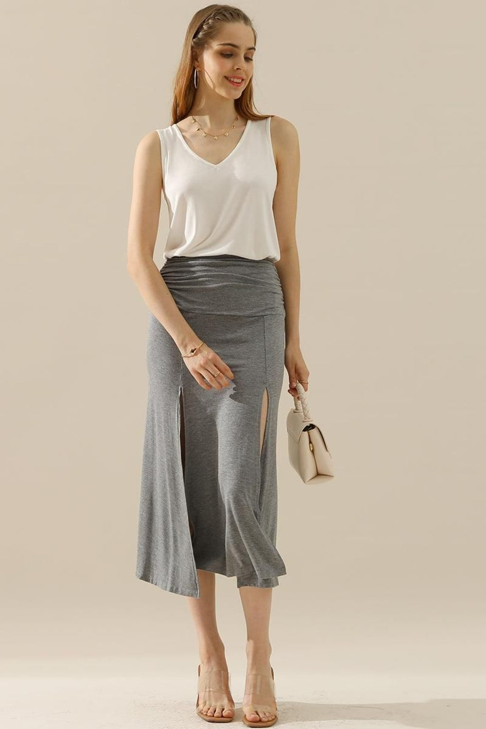 A woman in a stylish and versatile Ninexis Full Size V-Neck Curved Hem Tank and white pleated skirt poses while holding her handbag.