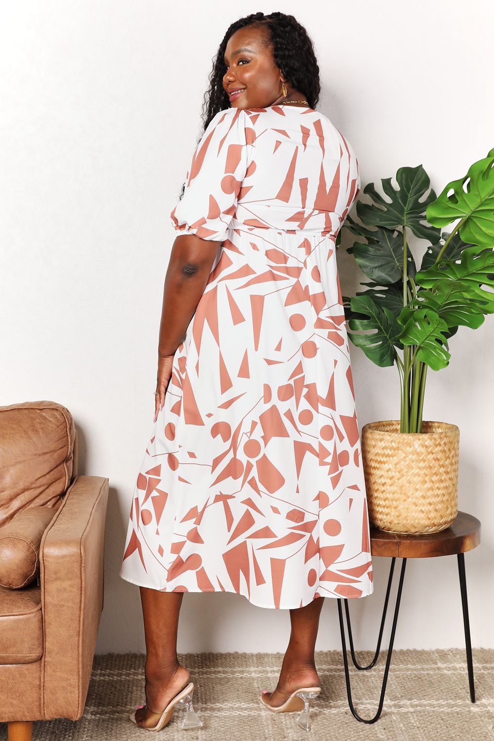 A person in a Printed Surplice Balloon Sleeve Dress stands near a brown chair and a round table with a potted plant.