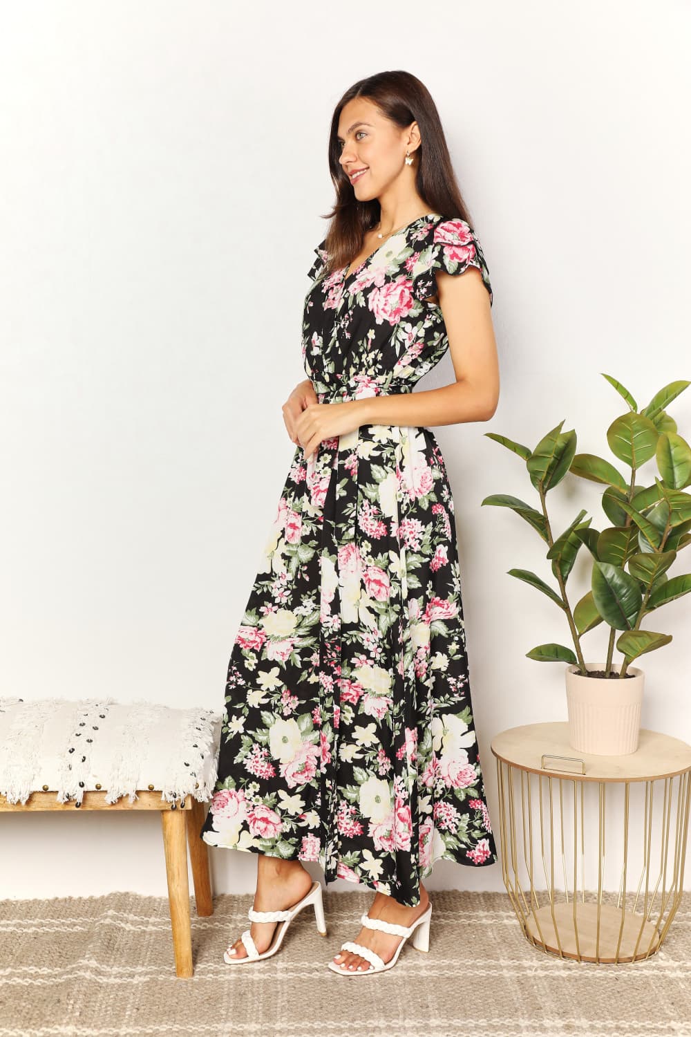 A woman wearing the Perfee Floral Flutter Sleeve Tie-Waist Split Dress stands beside a potted plant and a bench with textured fabric, perfect for adding charm to any vacation wardrobe.