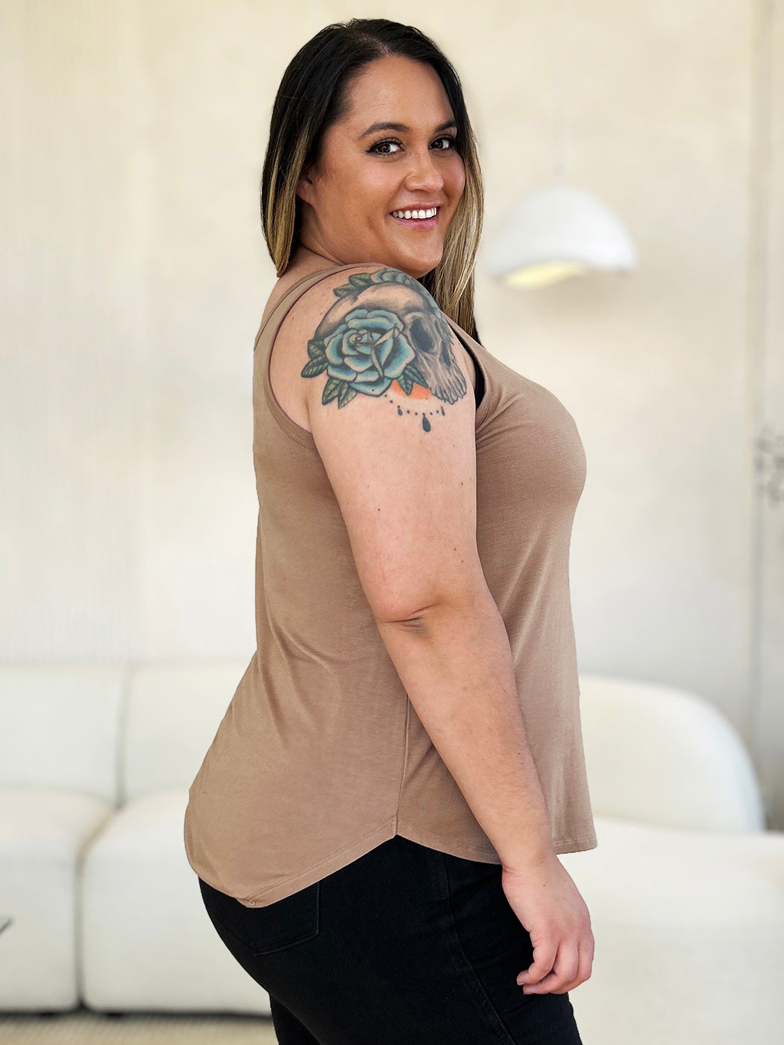 A woman wearing sunglasses, blue jeans, and the Basic Bae Full Size Round Neck Curved Hem Tank is standing in front of a concrete wall, holding her glasses with one hand and carrying a beige shoulder bag. Her basic style complements the slightly stretchy fabric of her outfit, ensuring comfort while maintaining chic simplicity.