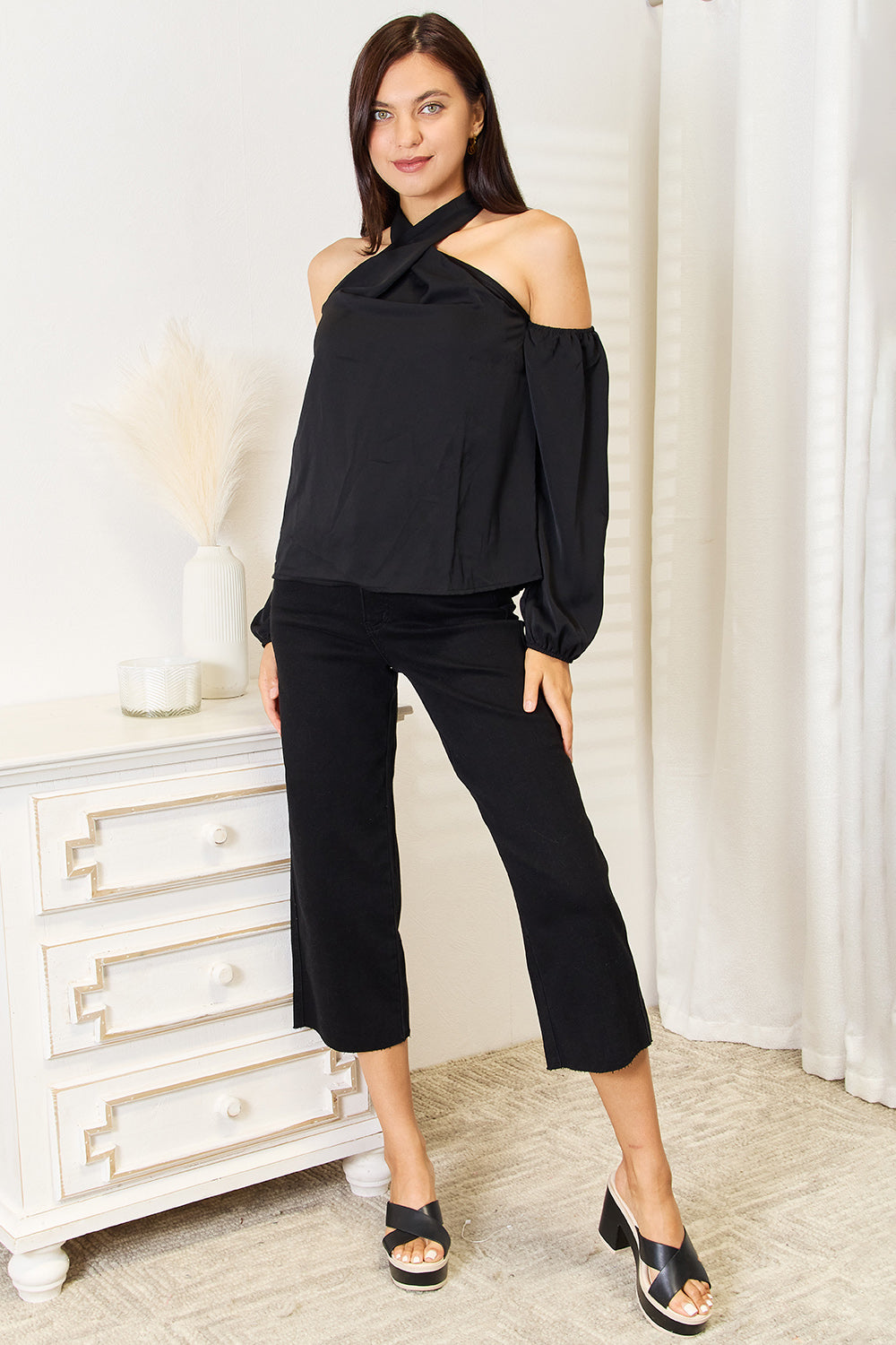 A woman with long brown hair wearing a Perfee Grecian Cold Shoulder Long Sleeve Blouse stands in a brightly lit room next to a white vase with pampas grass. She looks to the side with a smile.