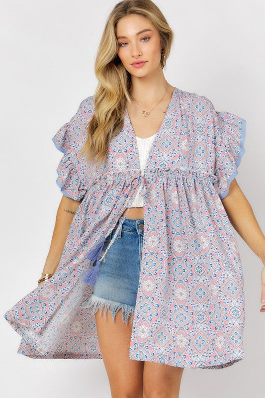 A woman stands against a white background wearing the Printed Short Sleeve Ruffle Kimono over a white top and denim shorts, effortlessly blending style and comfort.