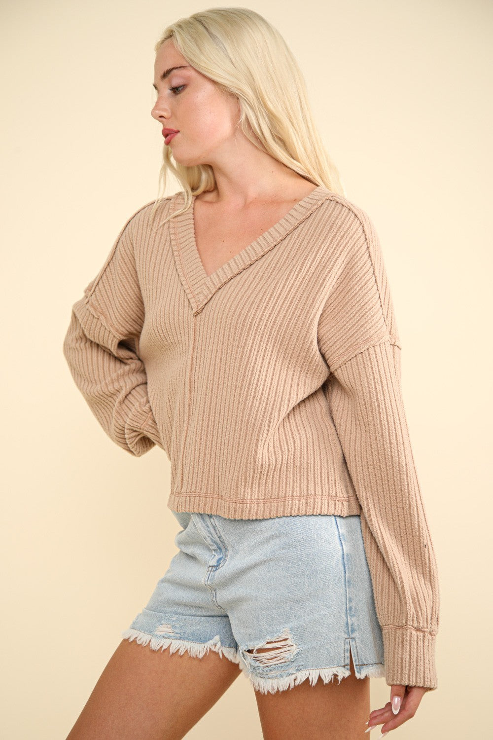 A woman with long blonde hair wears the VERY J Exposed Seam V-Neck Ribbed Knit Top, a casual and comfy piece featuring a ribbed knit texture and a drop shoulder design. She pairs it with light blue ripped denim shorts and stands against a cream-colored background.