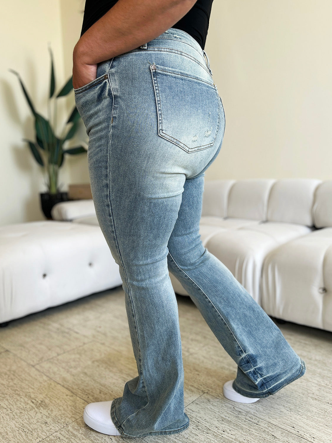 A person wearing Judy Blue Full Size Mid Rise Flare Jeans in a retro-inspired light blue color, paired with a white top and white shoes, stands in a modern living room with a white tufted sofa in the background.