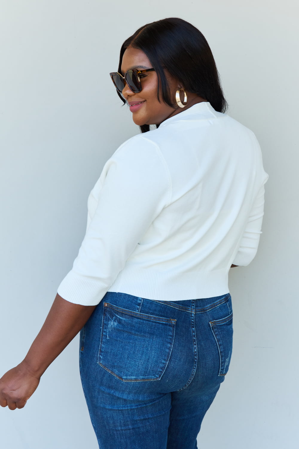 A stylish person wearing sunglasses, a Doublju My Favorite Full Size 3/4 Sleeve Cropped Cardigan in Ivory, and blue jeans poses in front of a plain background, effortlessly highlighting their versatile look.