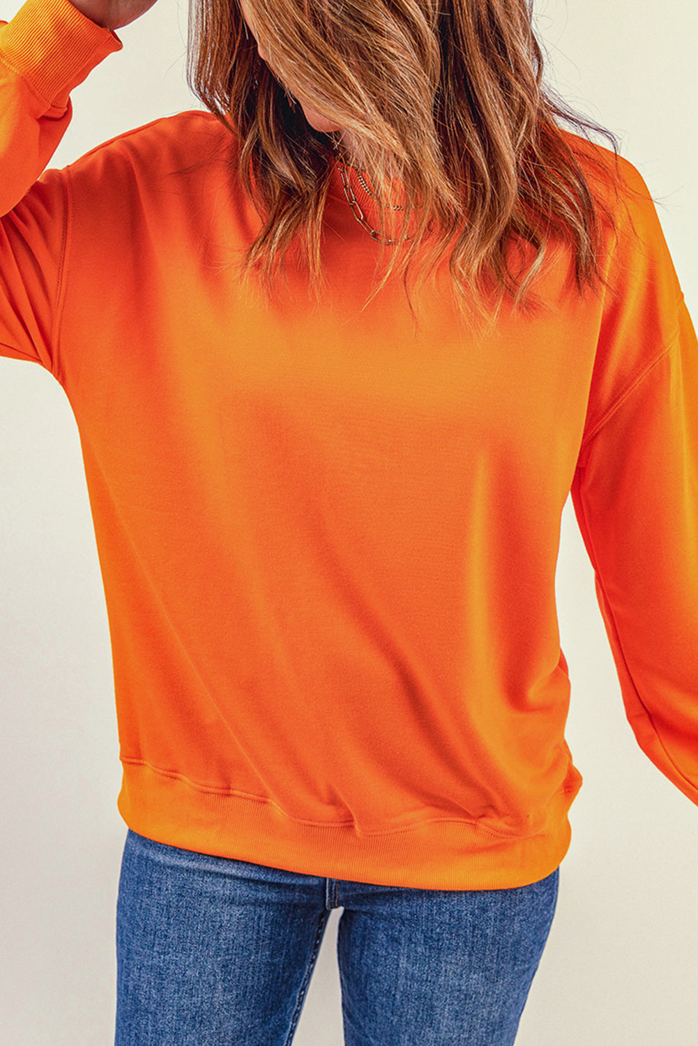 Person with wavy, shoulder-length hair wearing an Orange Plain Crew Neck Pullover Sweatshirt and blue jeans, standing with hand on hip, facing away.