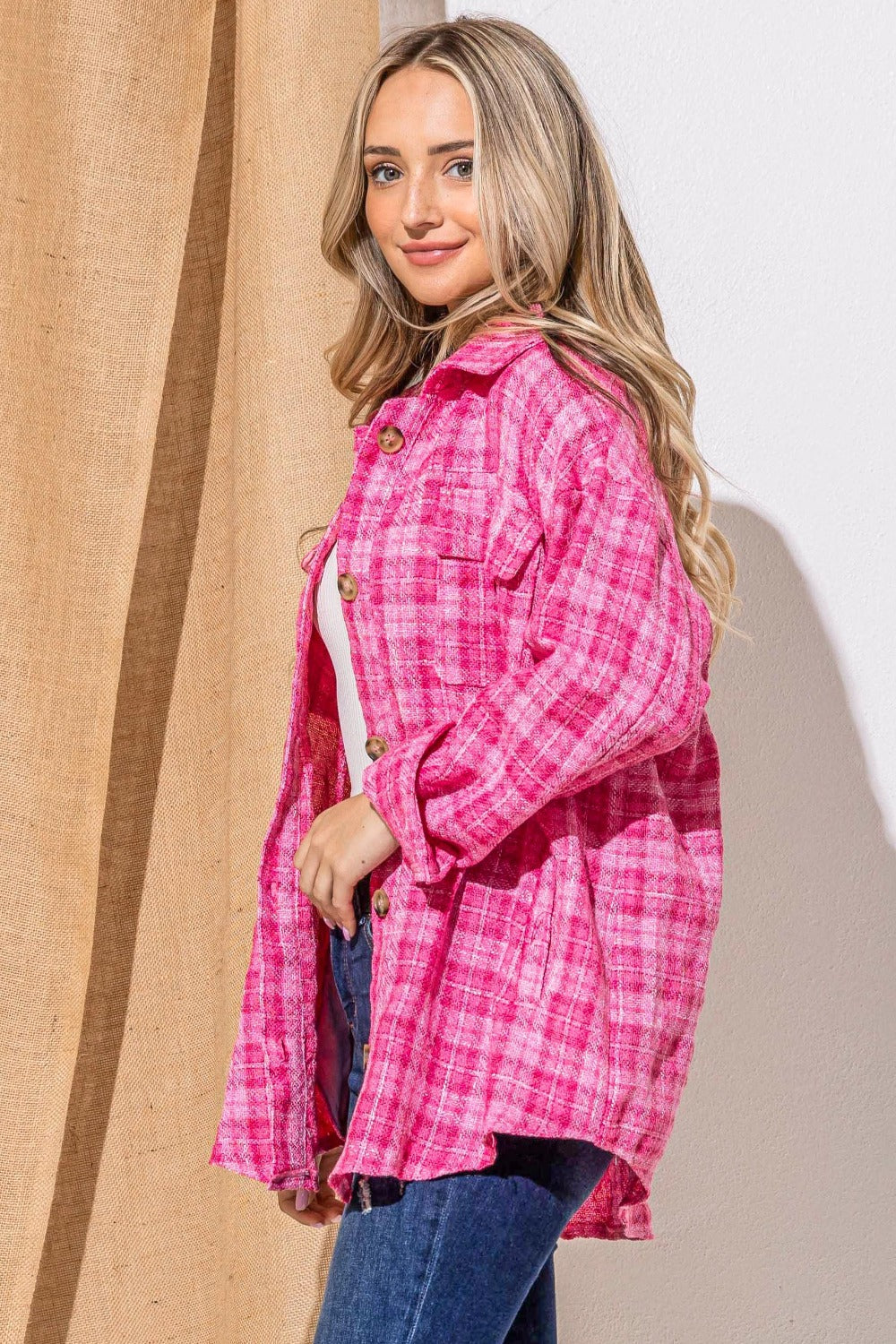 A woman stands smiling, wearing the "And The Why Full Size Tweed Button Down Long Sleeve Shacket" in pink plaid over a white top with a black belt and jeans. She is against a wall partially covered by a burlap curtain.