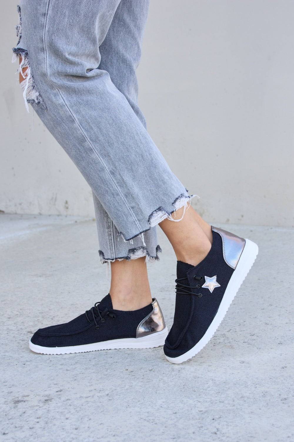 A person stands on a concrete surface, wearing distressed grey jeans and stylish Forever Link Star Lace-Up Flat Sneakers featuring black uppers and white soles.