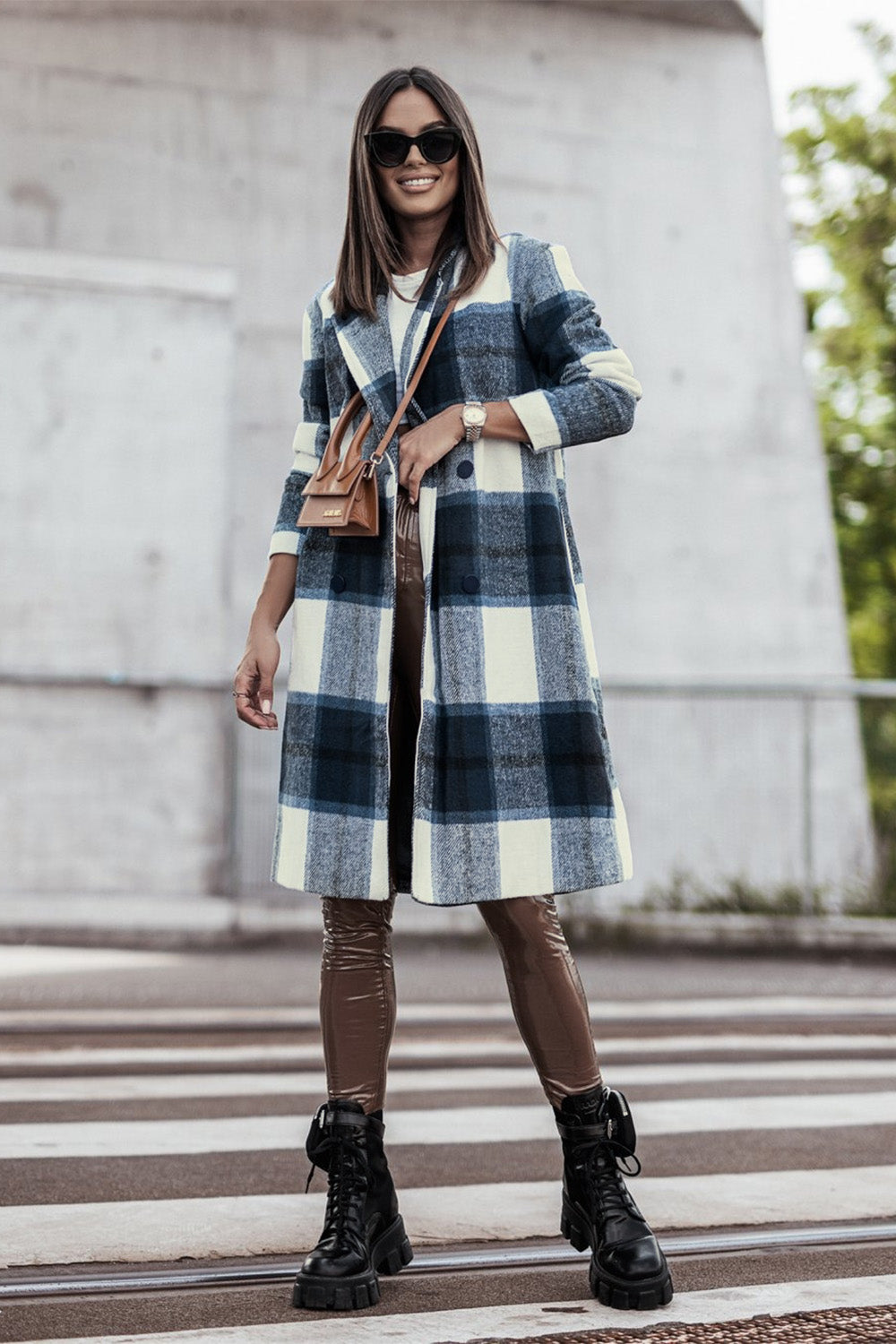 A woman stands on a sidewalk wearing the Double Take Full Size Plaid Button Up Lapel Collar Coat in pink and white over a white top and blue jeans, with her right hand lightly touching her hair. The polyester fabric of the coat makes it durable and perfect for an easy machine wash cold.