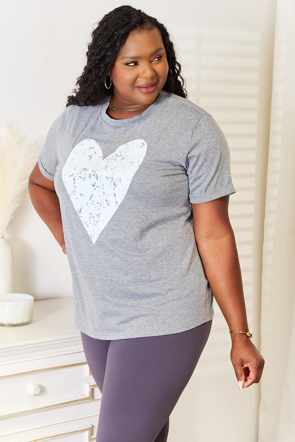 A woman with long hair stands indoors, smiling and touching her hair, wearing the trendy Simply Love Heart Graphic Cuffed Short Sleeve T-Shirt in gray.