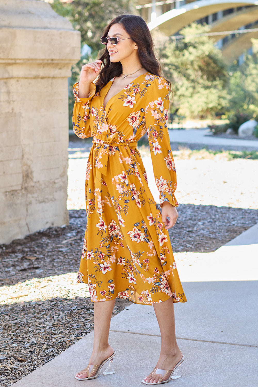 A woman wearing the Double Take Full Size Floral Tie Back Flounce Sleeve Dress in yellow, along with sunglasses, is walking on a paved path outdoors. She is smiling and has one hand on her hip.