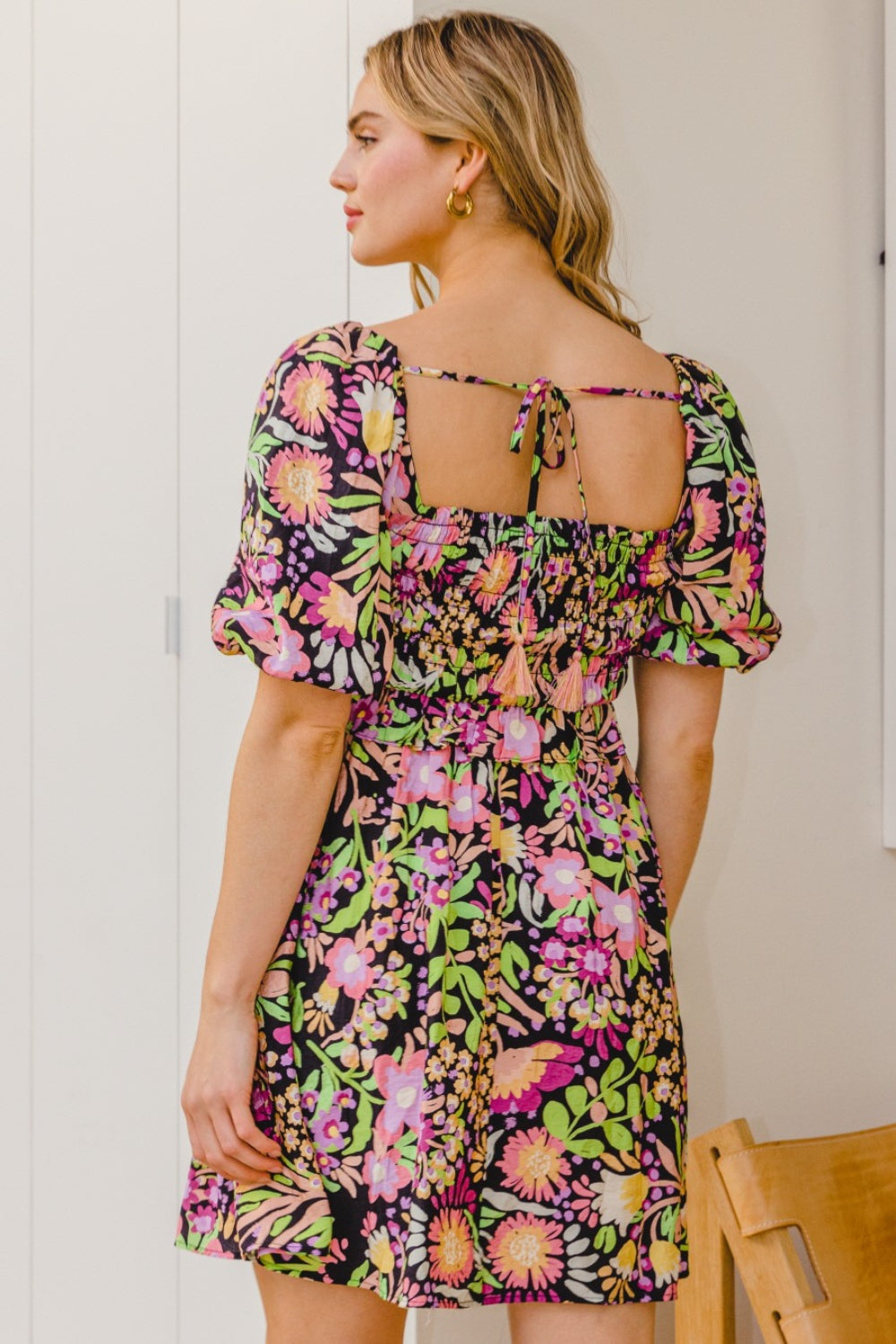 A person stands wearing the ODDI Full Size Floral Tie-Back Mini Dress, a feminine and flirty black floral design with puffy sleeves, perfect for a summer wedding.