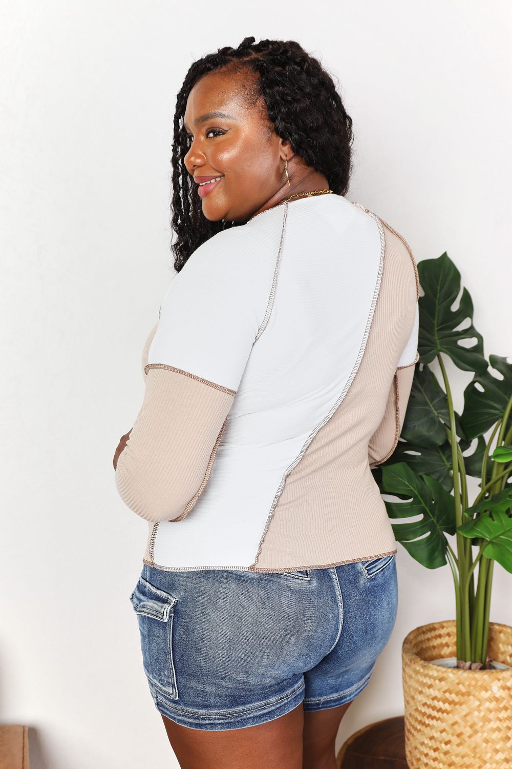 A woman with long brown hair is wearing the "Double Take Color Block Exposed Seam Top" in beige and white, paired with jeans. She stands with one hand in her pocket and the other touching her hair, smiling. A plant is visible in the background.