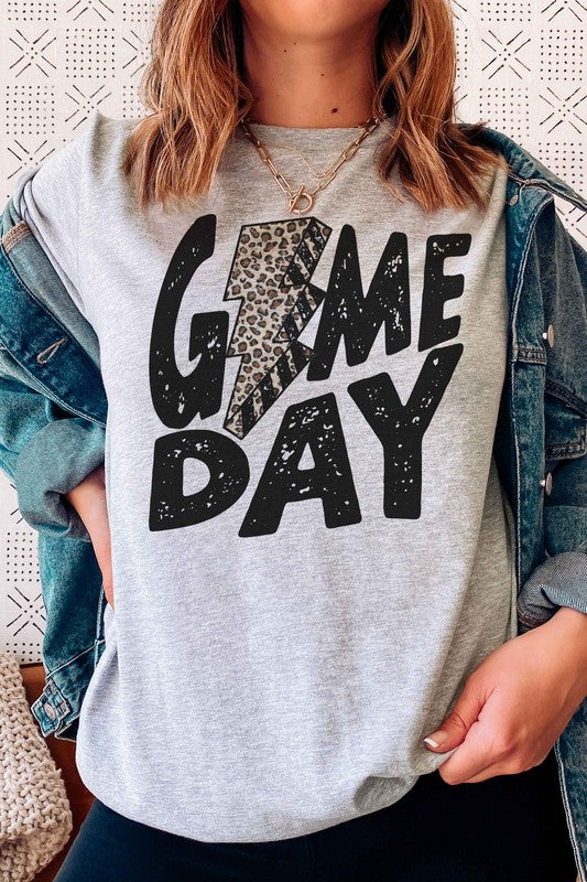 A person wearing a white "LEOPARD LIGHTNING GAME DAY Graphic Tee" with a leopard print lightning bolt design stands with a denim jacket partially on, showcasing its unisex sizing.