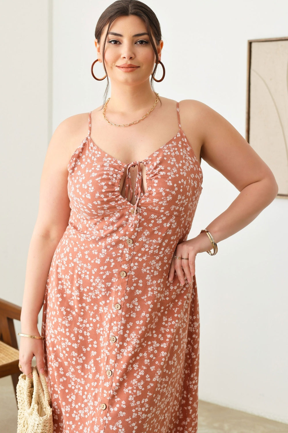 A woman stands indoors wearing the Zenobia Plus Size Cutout Floral Spaghetti Strap Dress in light blue, holding a small woven handbag. She is accessorized with earrings, bangles, and beige platform sandals.