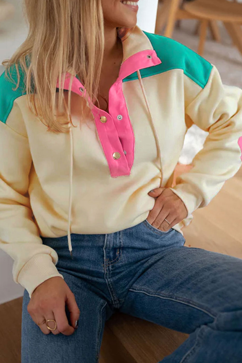 A woman wearing the Apricot Color Block Elbow Patch Half Button Sweatshirt, featuring cream and mint colors with pink elbow patches and a turn-down collar with snap button closure, stands outdoors. She has long wavy hair, sunglasses, a hat, and is wearing blue jeans.