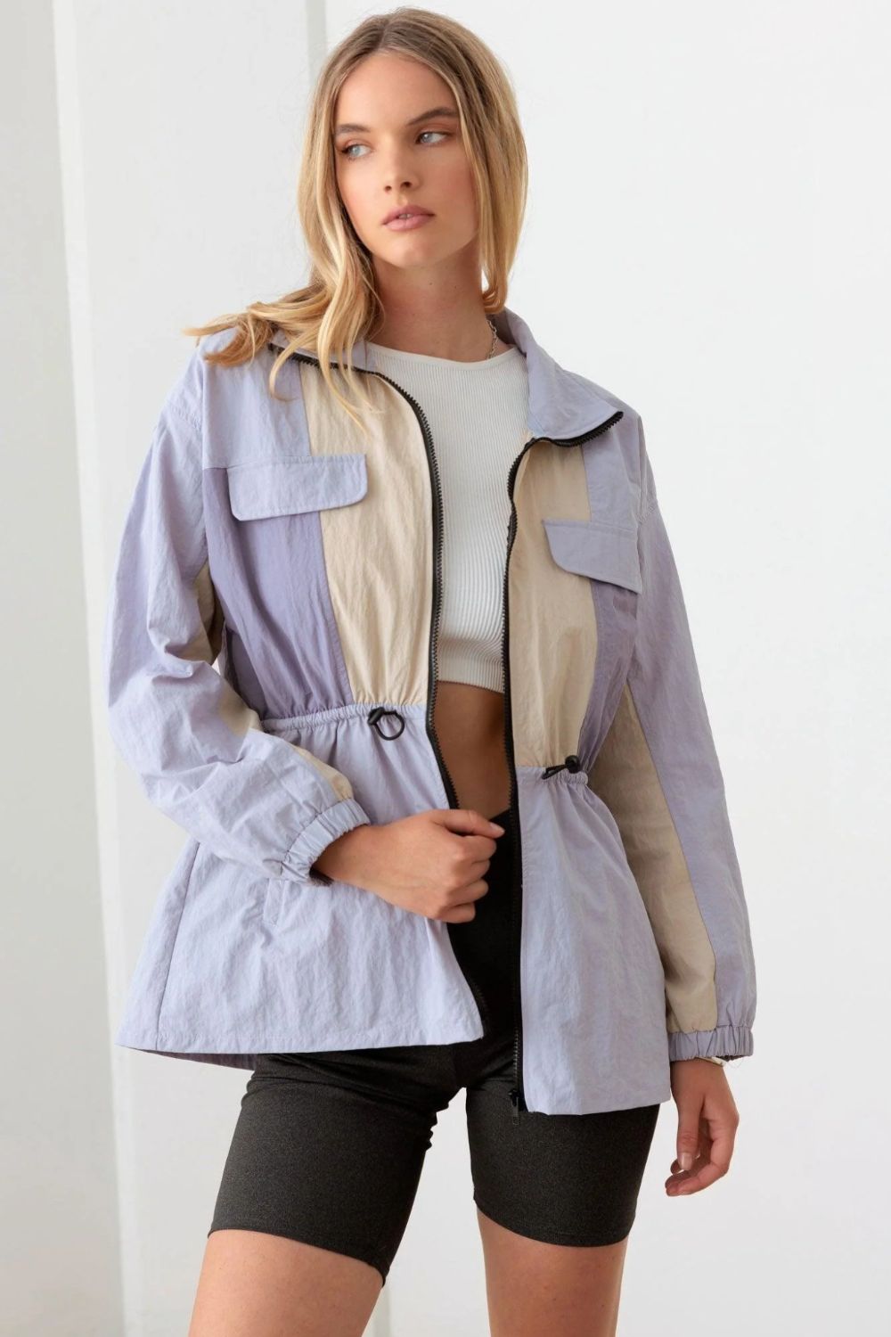 A woman wearing the Le Lis Color Block Collared Wind Breaker Jacket over a white crop top and black shorts stands in a well-lit room.