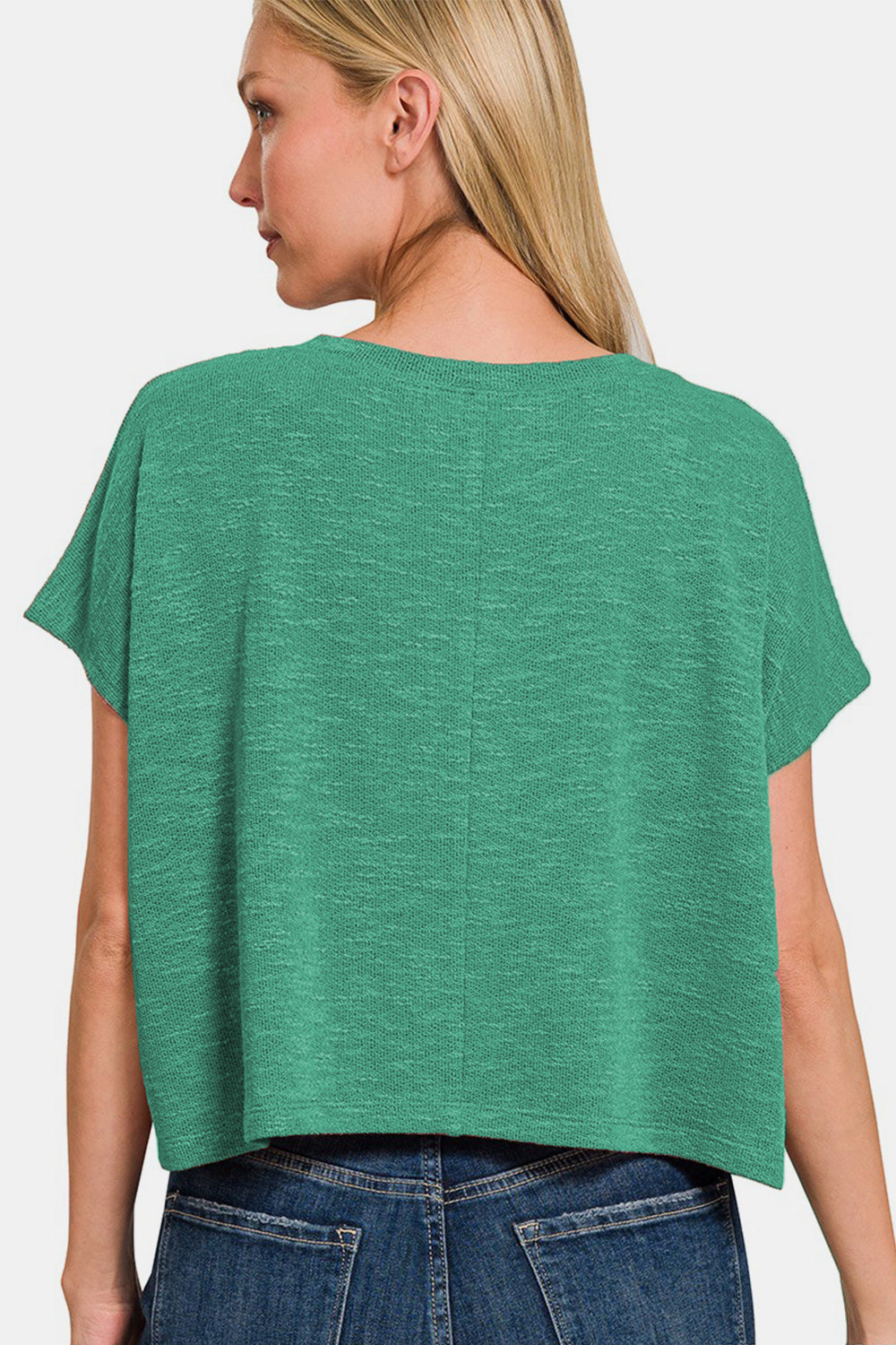 A person with long hair wears a green Zenana V-Neck Short Sleeve T-Shirt and blue jeans, standing against a white background. This versatile wardrobe essential offers both a stylish look and comfortable fit.