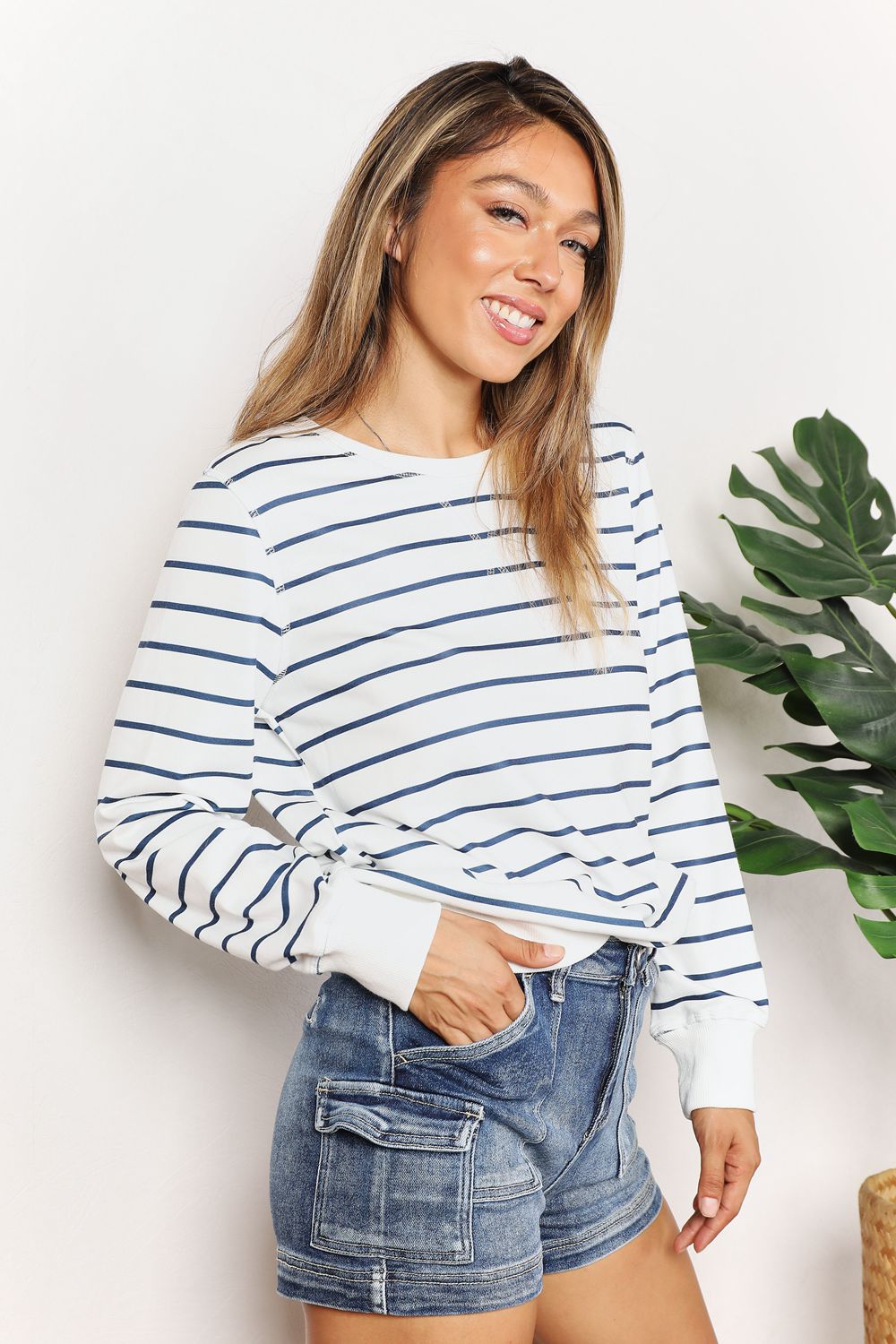 A person smiling while wearing the Double Take Striped Long Sleeve Round Neck Top, paired with denim shorts, stands in front of a white background with a plant.