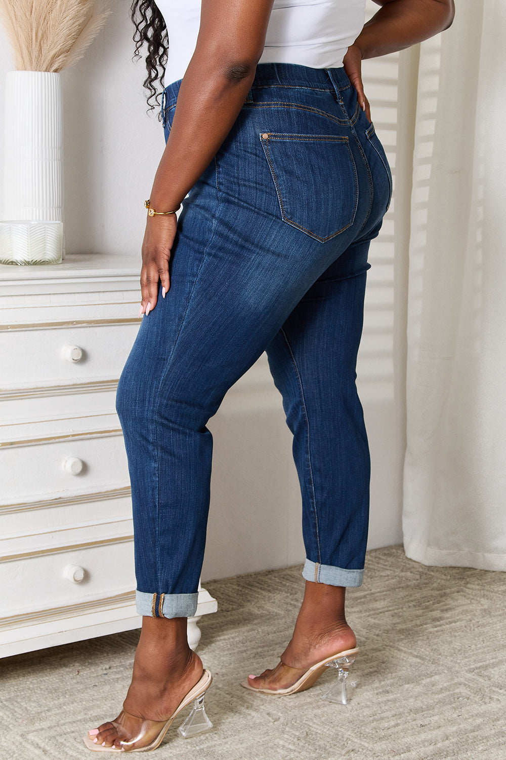 A person wearing Judy Blue Full Size Skinny Cropped Jeans with rolled-up cuffs and clear heels stands near a white dresser adorned with a decorative vase for a modern look.