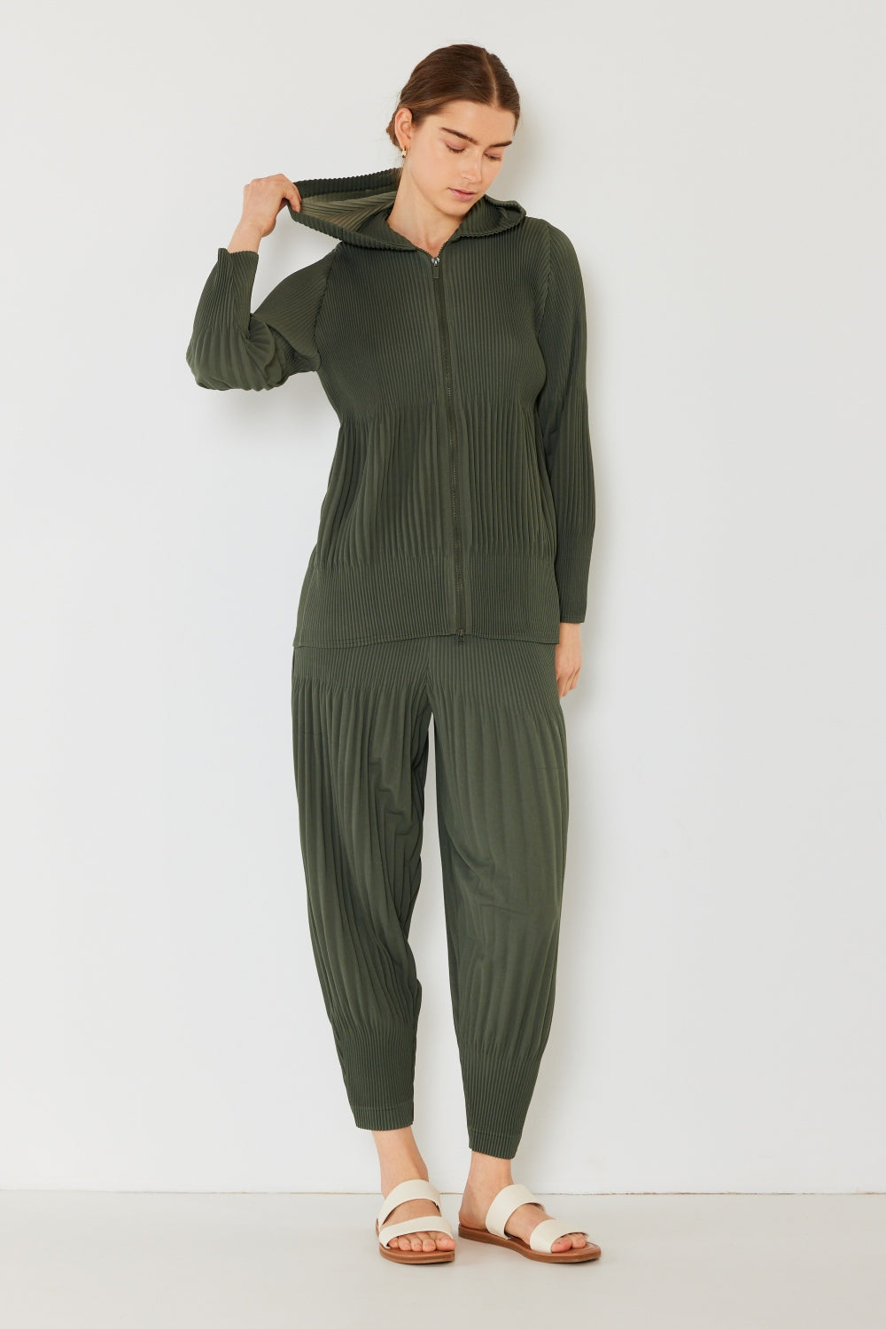 A person stands against a plain background, wearing a matching dark green hooded top and Marina West Swim Pleated Unisex Aladdin Plisse Pants with a ribbed texture, complemented by white sandals.