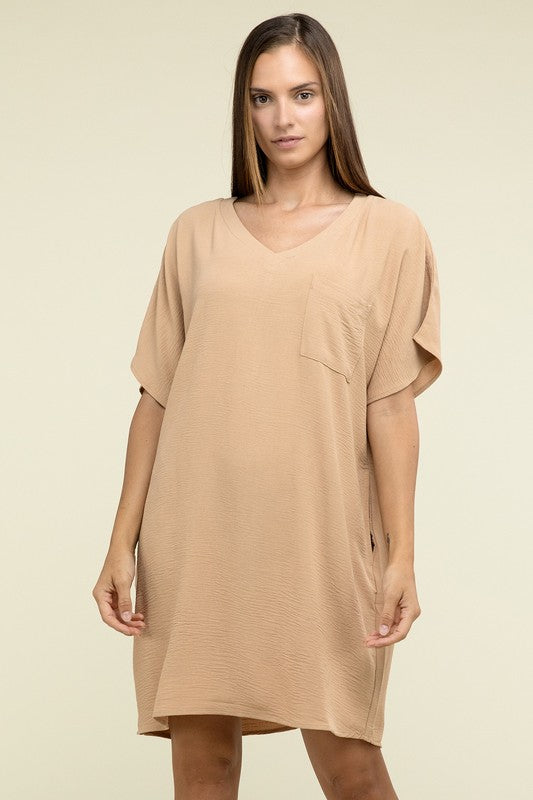 A woman wearing a loose light brown Woven Airflow V Neck T-Shirt Dress with Pockets stands against a plain background. Made from breathable fabric, this summer dress includes pockets where her hands rest as she smiles slightly.